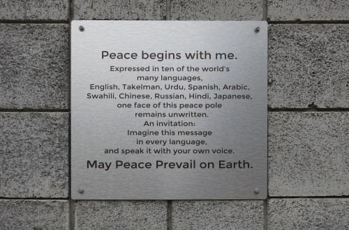 Metal plaque on brick wall of the church 'Peace begins with me. Expressed in ten of the world's many languages, English, Takelman, Urdu, Spanish, Arabic, Swahili, Chinese, Russian, Hindi, Japanese, one face of this peace pole remains unwritten. An invitation: Imagine this message in every language, and speak it with your own voice. May Peace Prevail On Earth.'